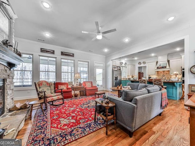 living area featuring light wood-type flooring, ceiling fan, crown molding, and a stone fireplace