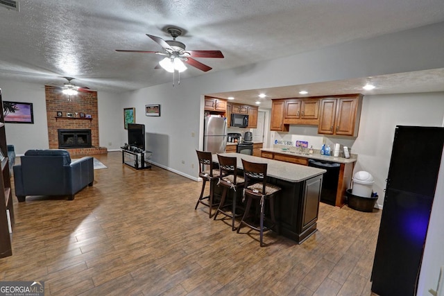 kitchen with a textured ceiling, wood finished floors, black appliances, brown cabinetry, and a kitchen bar