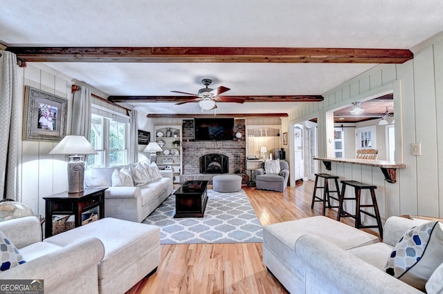 living area featuring wooden walls, a ceiling fan, light wood-style floors, a brick fireplace, and beamed ceiling