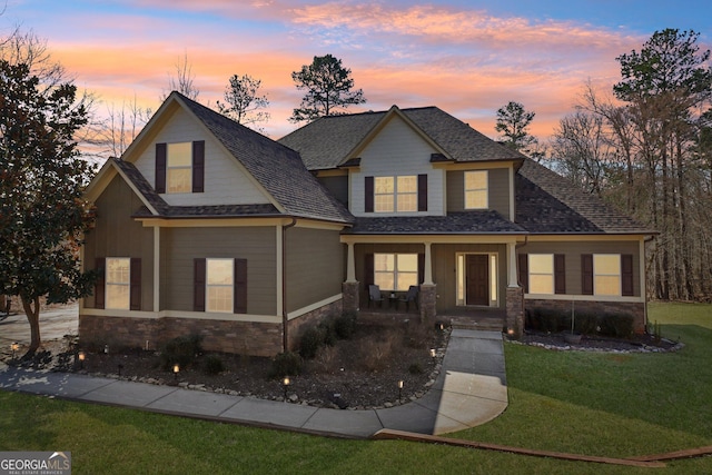 view of front facade with stone siding, covered porch, a yard, and roof with shingles