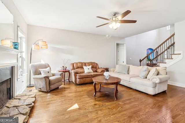 living room featuring light wood-type flooring, ceiling fan, stairs, and a fireplace with raised hearth