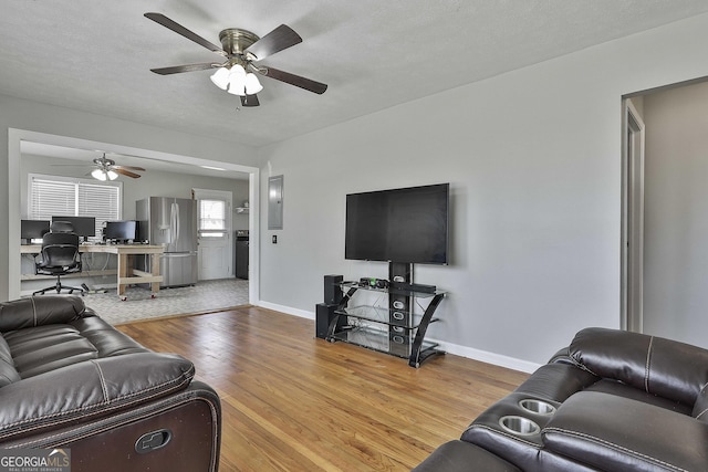 living room featuring a textured ceiling, light wood-style flooring, a ceiling fan, baseboards, and electric panel