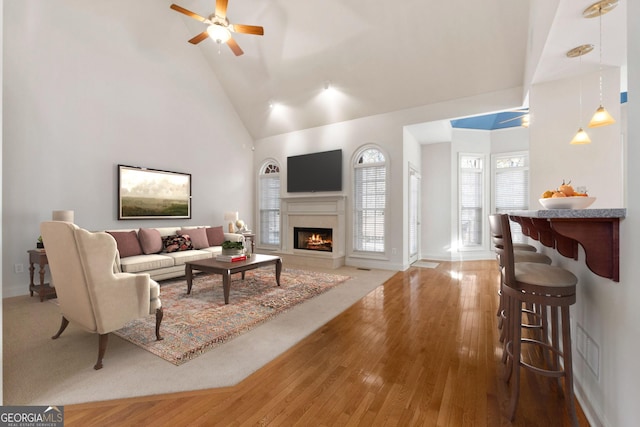 living area featuring wood-type flooring, ceiling fan, high vaulted ceiling, a lit fireplace, and baseboards