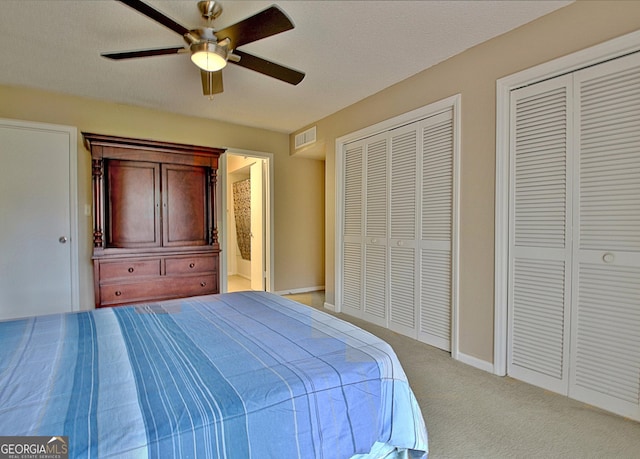 bedroom with ceiling fan, ensuite bathroom, light colored carpet, visible vents, and two closets