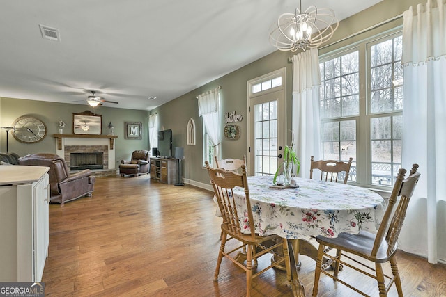 dining area with visible vents, a fireplace with raised hearth, ceiling fan with notable chandelier, light wood finished floors, and baseboards