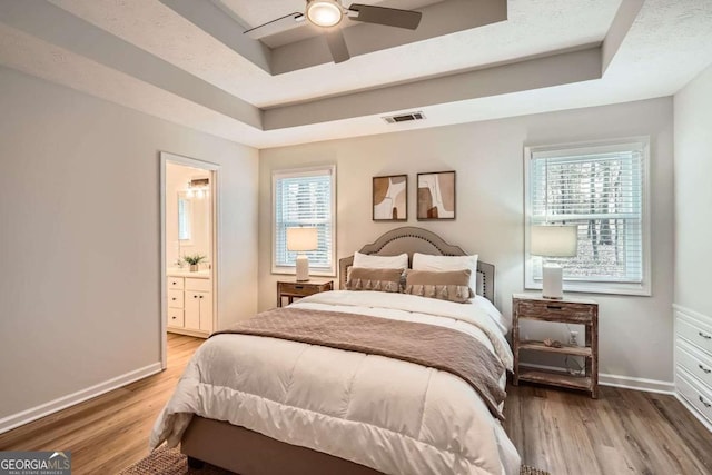 bedroom with baseboards, visible vents, a raised ceiling, wood finished floors, and ensuite bathroom