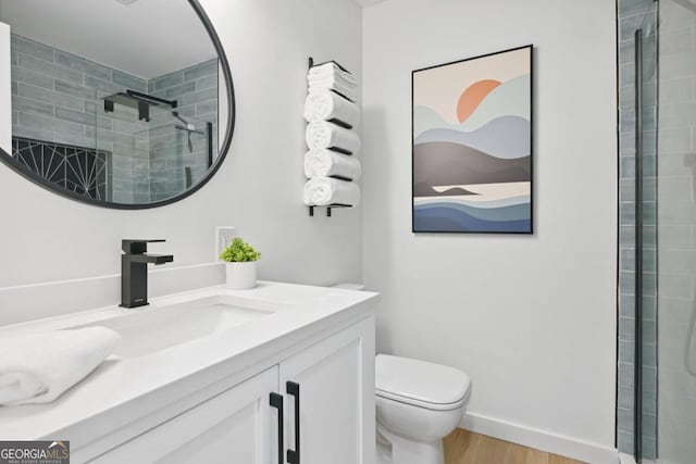 bathroom with vanity, wood finished floors, baseboards, tiled shower, and toilet