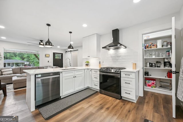 kitchen with a sink, open floor plan, a peninsula, appliances with stainless steel finishes, and wall chimney exhaust hood