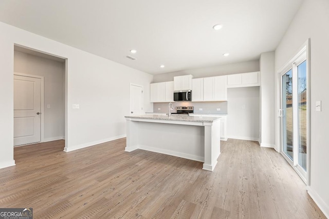 kitchen featuring baseboards, an island with sink, stainless steel appliances, light wood-type flooring, and white cabinetry