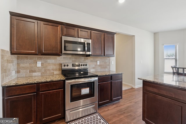 kitchen featuring light stone countertops, light wood-style flooring, dark brown cabinets, appliances with stainless steel finishes, and backsplash