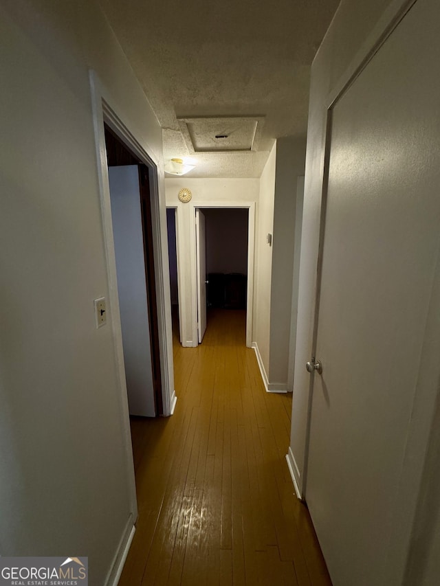 hallway featuring a textured ceiling, hardwood / wood-style flooring, attic access, and baseboards