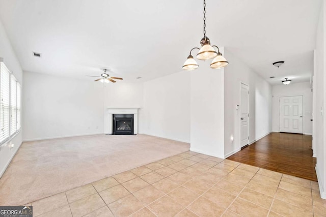 unfurnished living room with light colored carpet, visible vents, a fireplace with flush hearth, light tile patterned flooring, and ceiling fan with notable chandelier