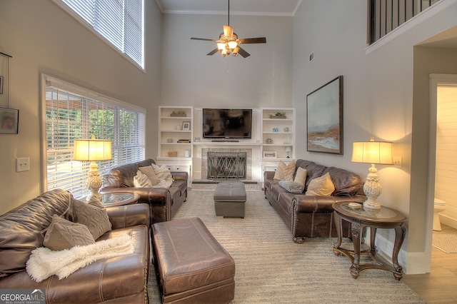 living area featuring a fireplace with raised hearth, a ceiling fan, a towering ceiling, ornamental molding, and wood finished floors