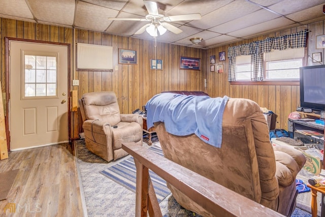 living room with wooden walls, a ceiling fan, a drop ceiling, and wood finished floors