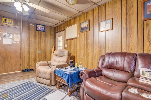 living area featuring a drop ceiling, ceiling fan, wooden walls, and wood finished floors