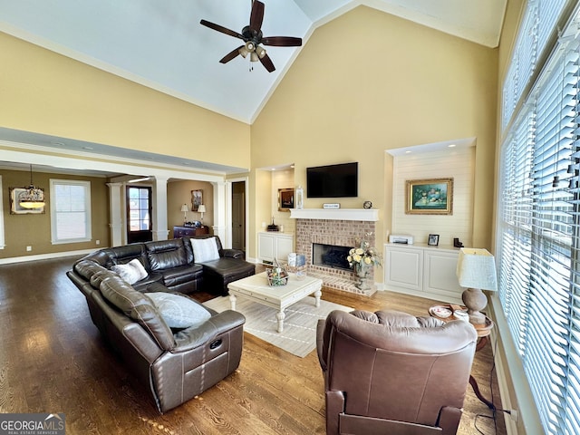 living room with ceiling fan, high vaulted ceiling, wood finished floors, a brick fireplace, and decorative columns