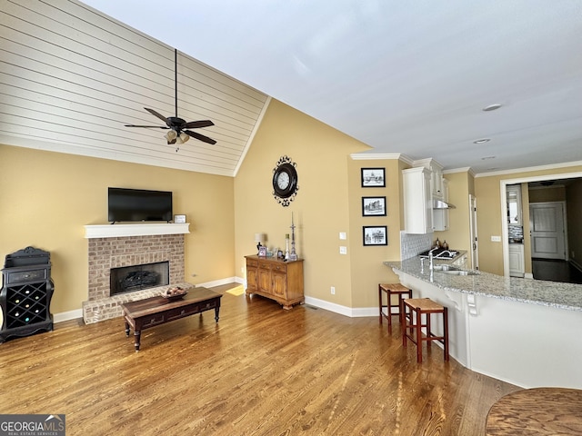 living area with wood finished floors, baseboards, vaulted ceiling, ornamental molding, and a brick fireplace