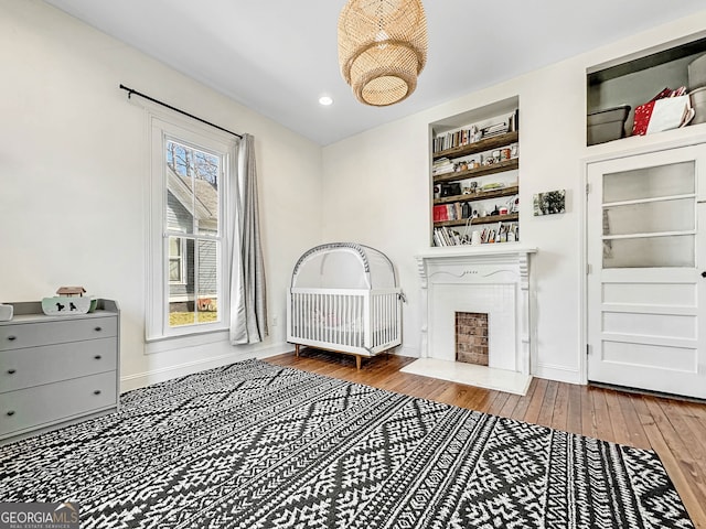 bedroom featuring recessed lighting, a fireplace with flush hearth, a nursery area, baseboards, and hardwood / wood-style flooring