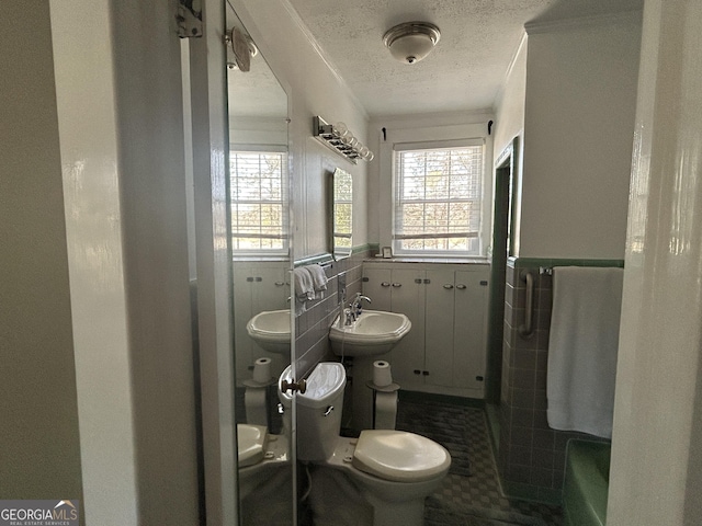 full bath featuring toilet, a sink, a textured ceiling, tile walls, and wainscoting