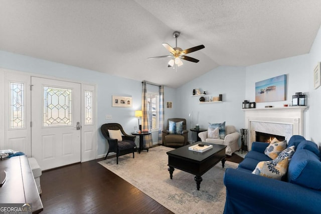 living area with ceiling fan, a textured ceiling, dark wood-type flooring, a high end fireplace, and vaulted ceiling