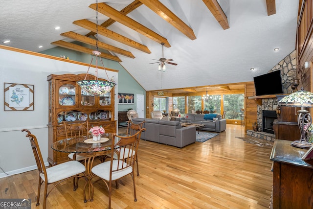 dining room featuring light wood-type flooring, beamed ceiling, high vaulted ceiling, ceiling fan with notable chandelier, and a stone fireplace