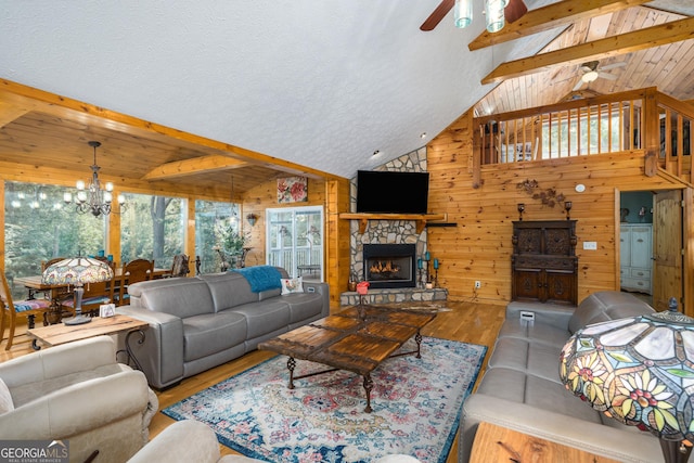 living room featuring wood finished floors, vaulted ceiling with beams, a stone fireplace, wood walls, and ceiling fan with notable chandelier