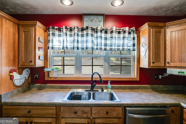 kitchen featuring a textured ceiling, stainless steel dishwasher, brown cabinetry, and a sink