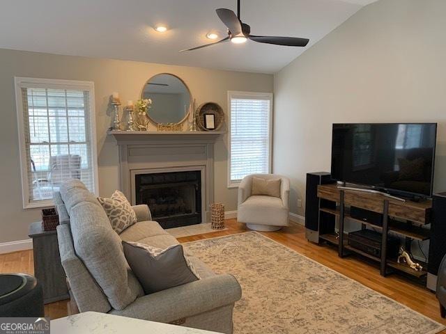 living area featuring baseboards, a fireplace with flush hearth, vaulted ceiling, wood finished floors, and a ceiling fan