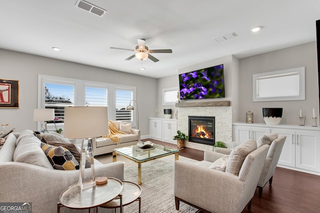 living room with dark wood-type flooring, visible vents, a stone fireplace, and a ceiling fan