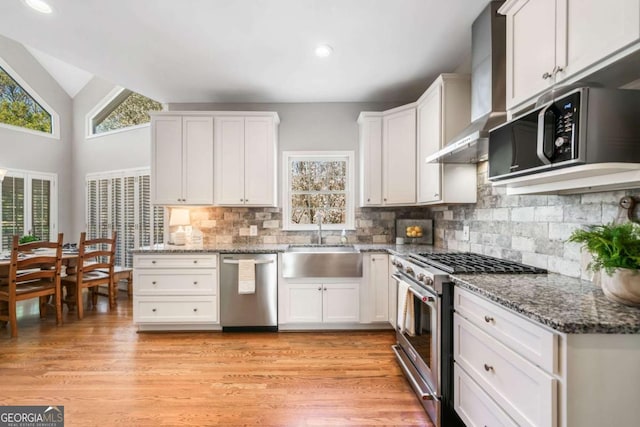 kitchen featuring lofted ceiling, a sink, white cabinets, appliances with stainless steel finishes, and wall chimney exhaust hood