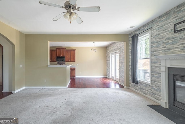 unfurnished living room featuring arched walkways, dark carpet, a large fireplace, baseboards, and ceiling fan with notable chandelier