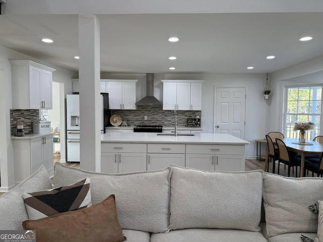 kitchen featuring white refrigerator with ice dispenser, open floor plan, white cabinetry, a sink, and wall chimney exhaust hood