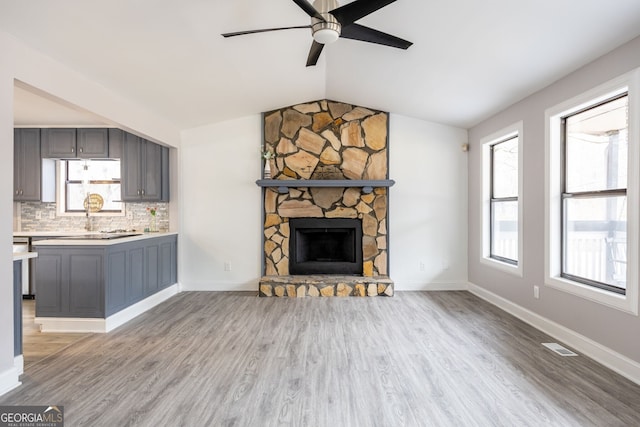 unfurnished living room with baseboards, ceiling fan, vaulted ceiling, a stone fireplace, and light wood-type flooring