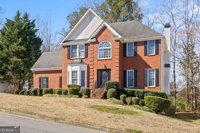 colonial-style house featuring brick siding, a chimney, a shingled roof, and a front lawn