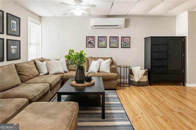 living area with baseboards, a ceiling fan, a wall unit AC, light wood-style flooring, and a paneled ceiling