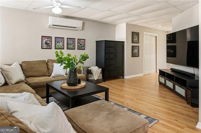 living area featuring light wood finished floors, baseboards, a ceiling fan, a drop ceiling, and a wall mounted air conditioner