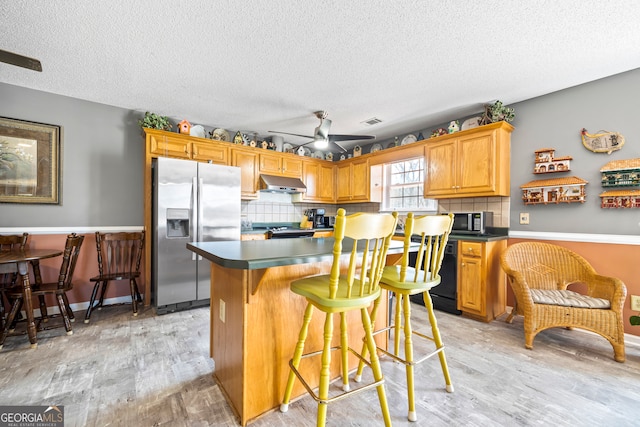 kitchen featuring dark countertops, a kitchen island, under cabinet range hood, light wood-style flooring, and stainless steel appliances