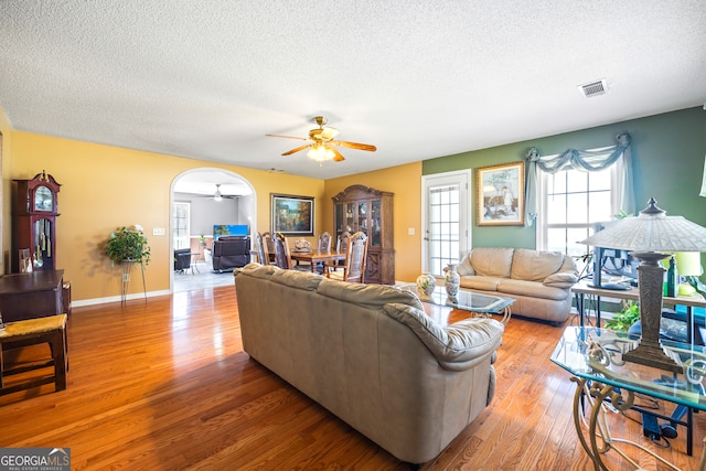 living room with visible vents, light wood-style floors, arched walkways, a textured ceiling, and a ceiling fan