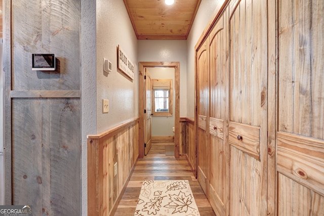 hall with a wainscoted wall, light wood-style floors, wooden walls, wooden ceiling, and a textured wall