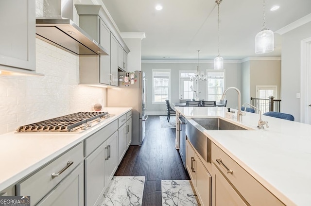 kitchen with light countertops, ornamental molding, stainless steel appliances, wall chimney exhaust hood, and a sink