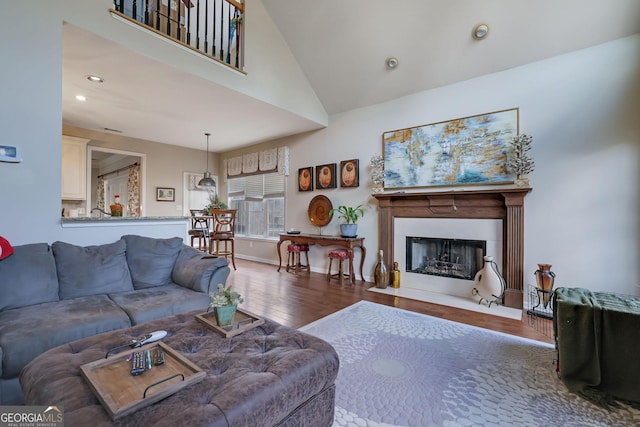 living area with wood finished floors, baseboards, a fireplace with flush hearth, recessed lighting, and a towering ceiling