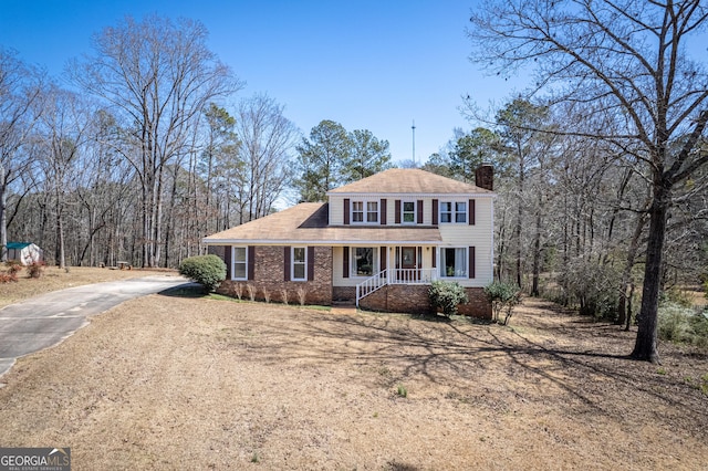 view of front facade featuring brick siding, dirt driveway, covered porch, a view of trees, and a chimney