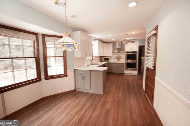 kitchen featuring a sink, dark wood finished floors, stainless steel double oven, wall chimney exhaust hood, and light countertops