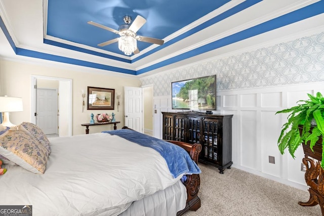 carpeted bedroom featuring wainscoting, a decorative wall, a ceiling fan, a tray ceiling, and ornamental molding