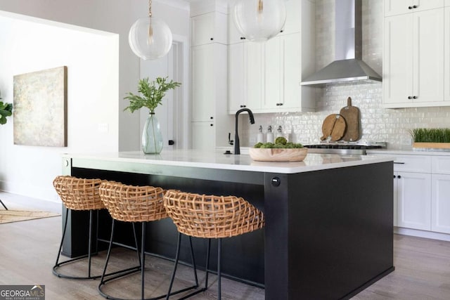 interior space with light wood-type flooring, a center island with sink, backsplash, wall chimney exhaust hood, and white cabinets