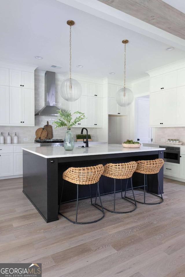 kitchen with light wood-style flooring, tasteful backsplash, white cabinetry, wall chimney range hood, and a large island with sink