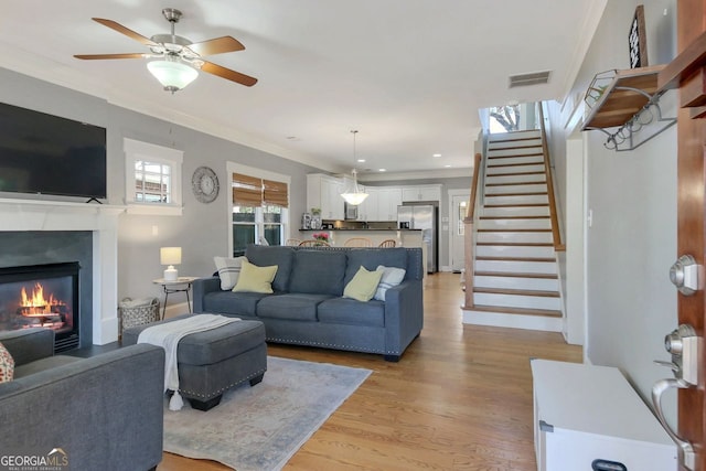 living room with visible vents, ornamental molding, light wood-style flooring, a glass covered fireplace, and stairs