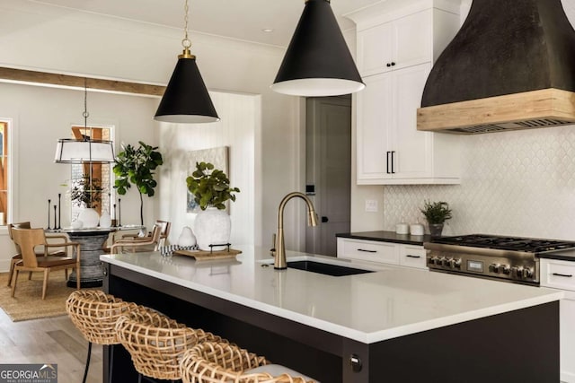 kitchen featuring an island with sink, light wood-style flooring, custom range hood, a sink, and decorative backsplash