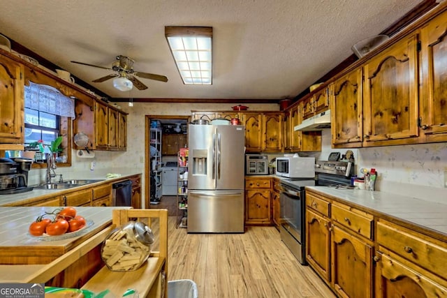 kitchen featuring under cabinet range hood, tile countertops, light wood-style floors, appliances with stainless steel finishes, and brown cabinetry