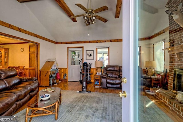 living room featuring a wainscoted wall, a ceiling fan, lofted ceiling with beams, wood finished floors, and a brick fireplace